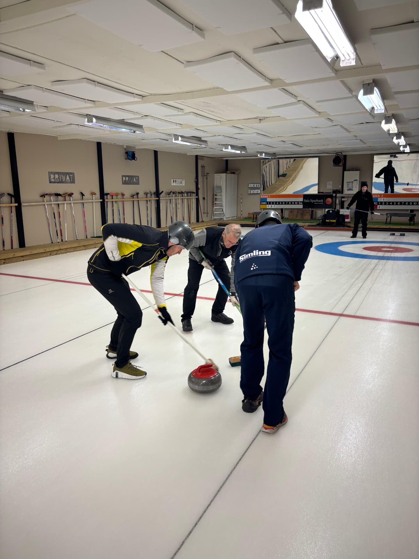 Tre kollegor sopar framför en curlingsten i curlinghallen