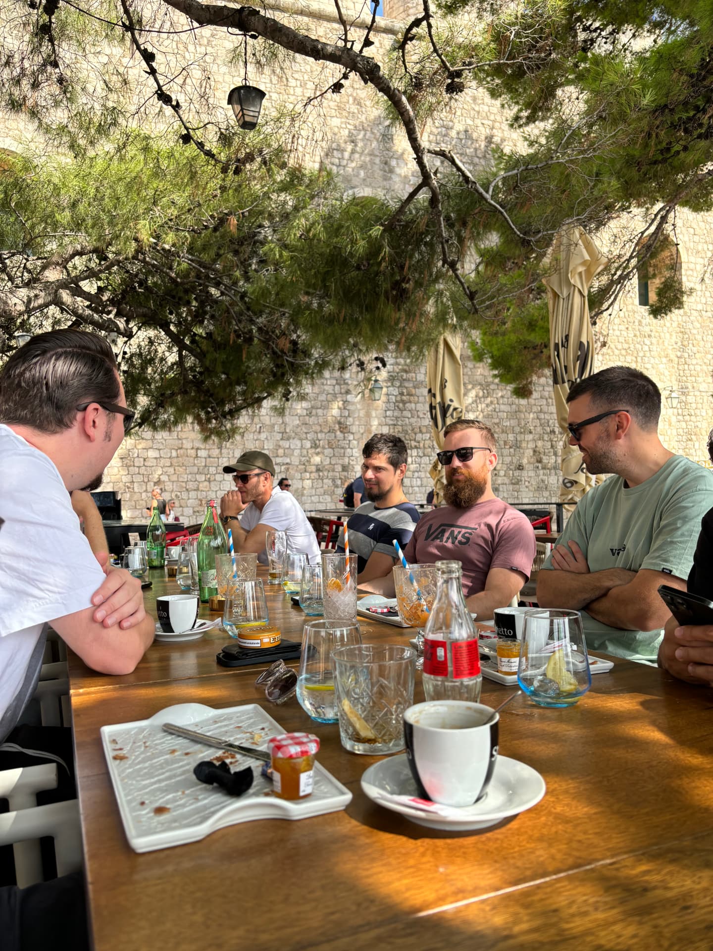 Stort sällskap runt ett träbord under ett pinjeträd vid en stenmur i Dubrovnik